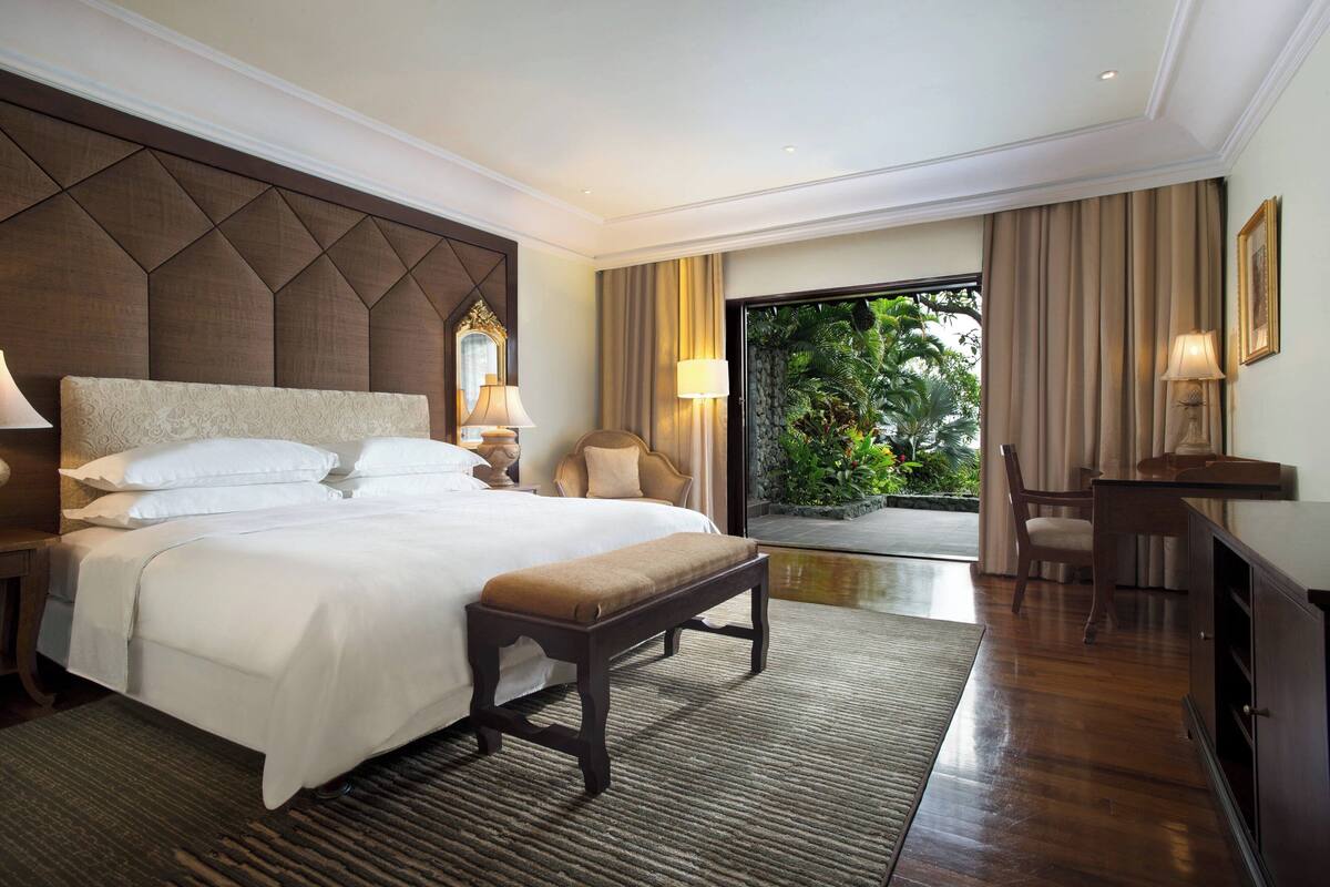A hotel room with a large bed, a bench, a desk, and a view of greenery through the glass door.