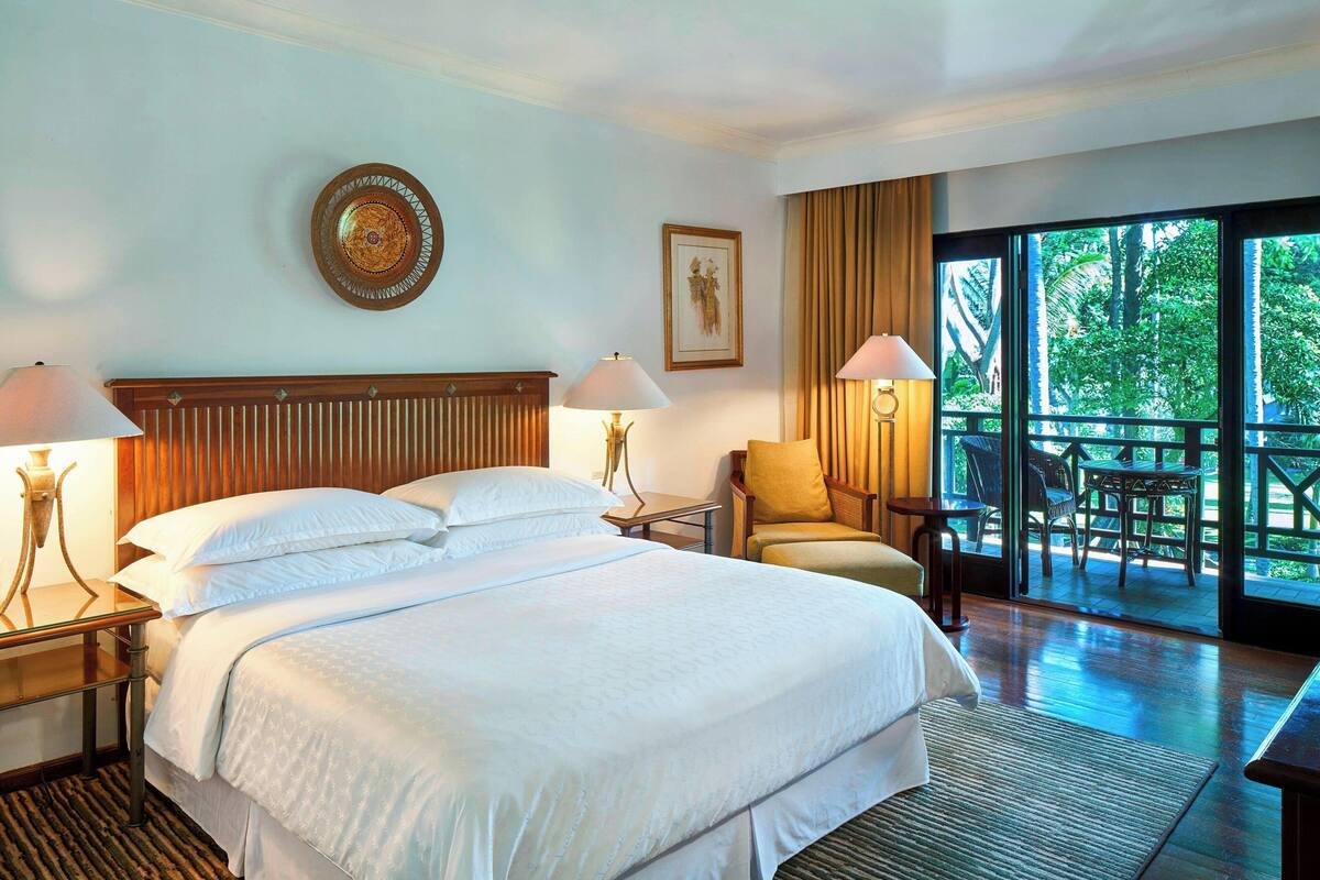 A hotel room with a large bed, two bedside tables with lamps, a wooden headboard, a framed picture on the wall, and a balcony with outdoor furniture.