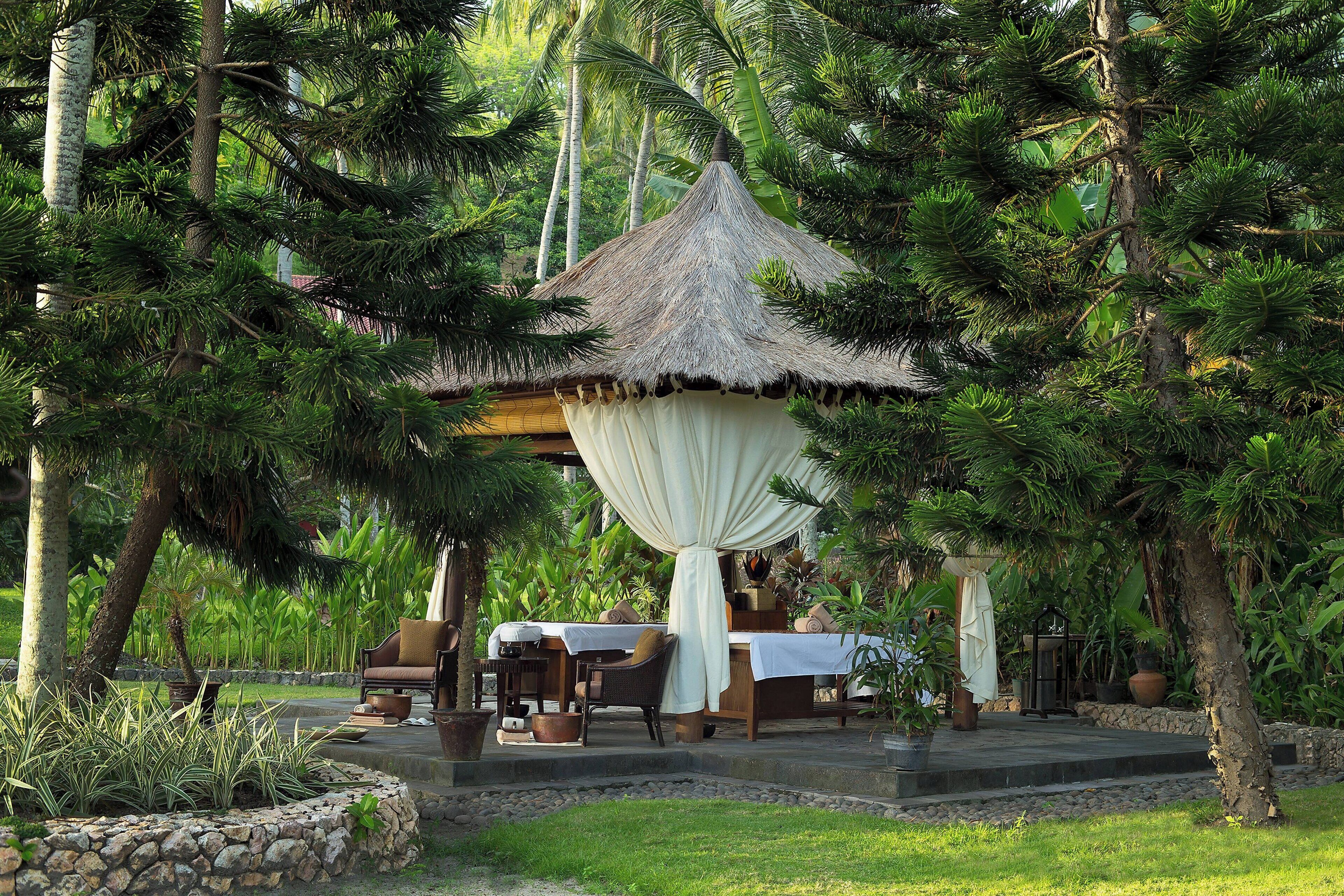 A thatched-roof gazebo with a white canopy, surrounded by lush greenery and a stone wall.