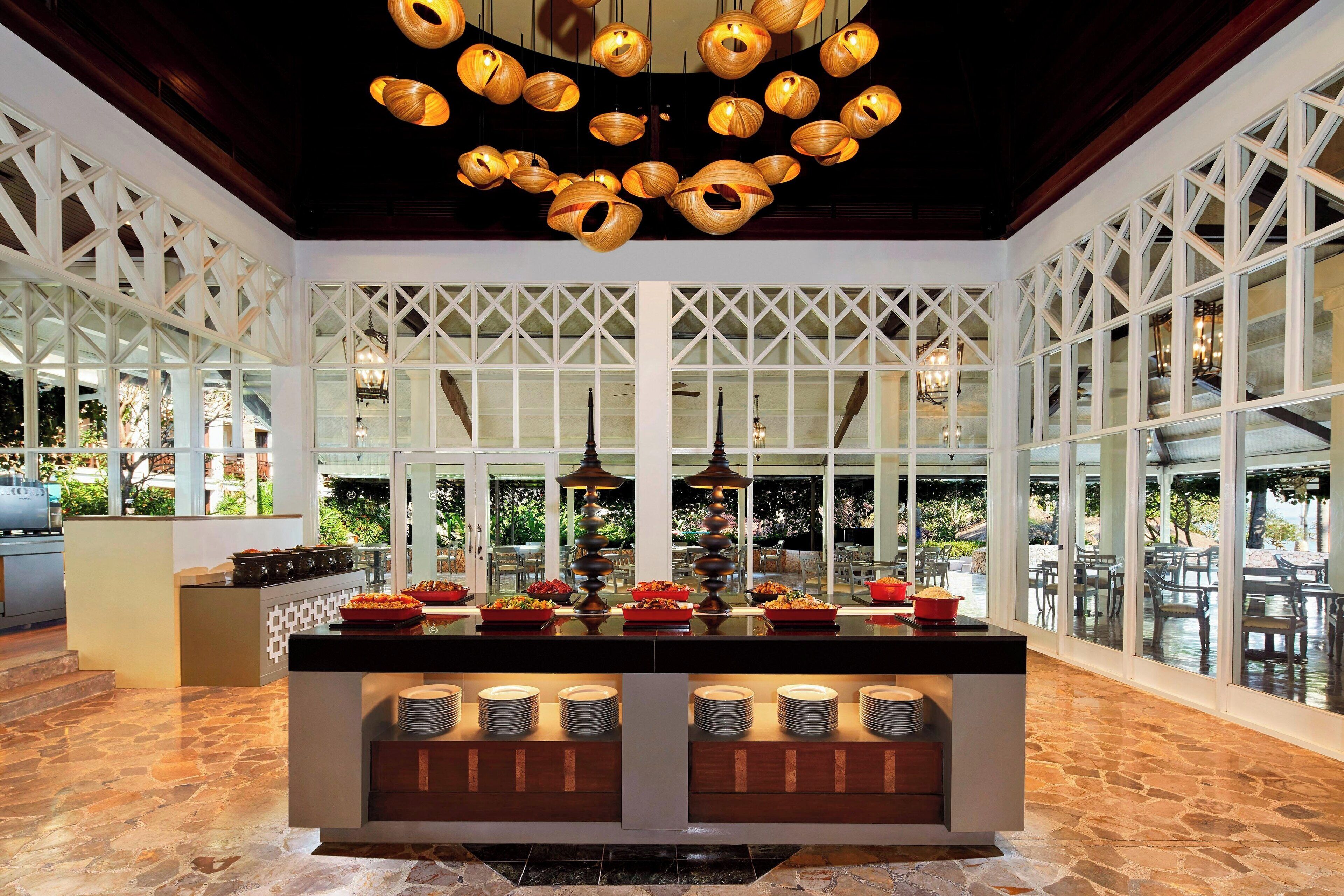 A buffet area with a central island, overhead wooden chandeliers, and large windows offering views of the outdoors.