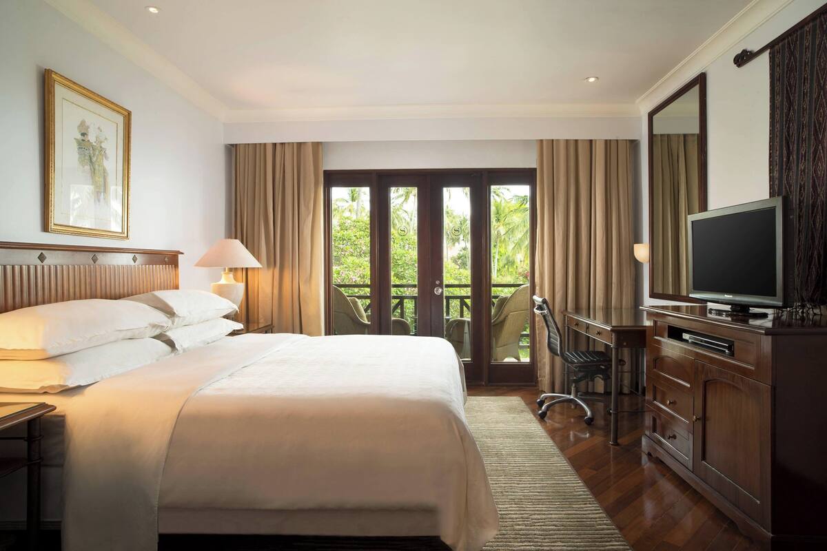 A hotel room with a large bed, a desk with a chair, a television, and a balcony with curtains.