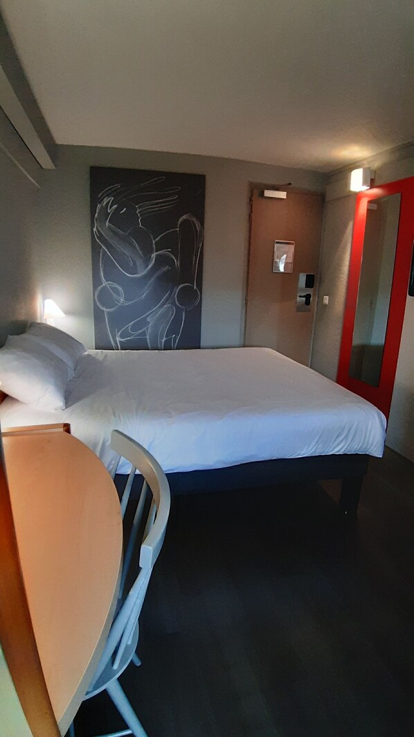 A hotel room with a bed, a desk, a chair, a wall with a chalkboard, and a red door.