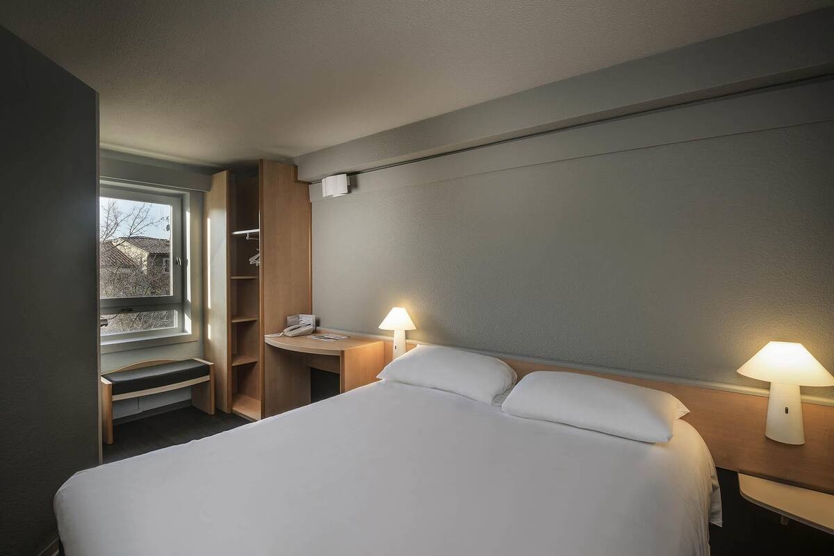 Une chambre d’hôtel avec un lit, des lampes de chevet, un bureau avec un téléphone et une fenêtre.
