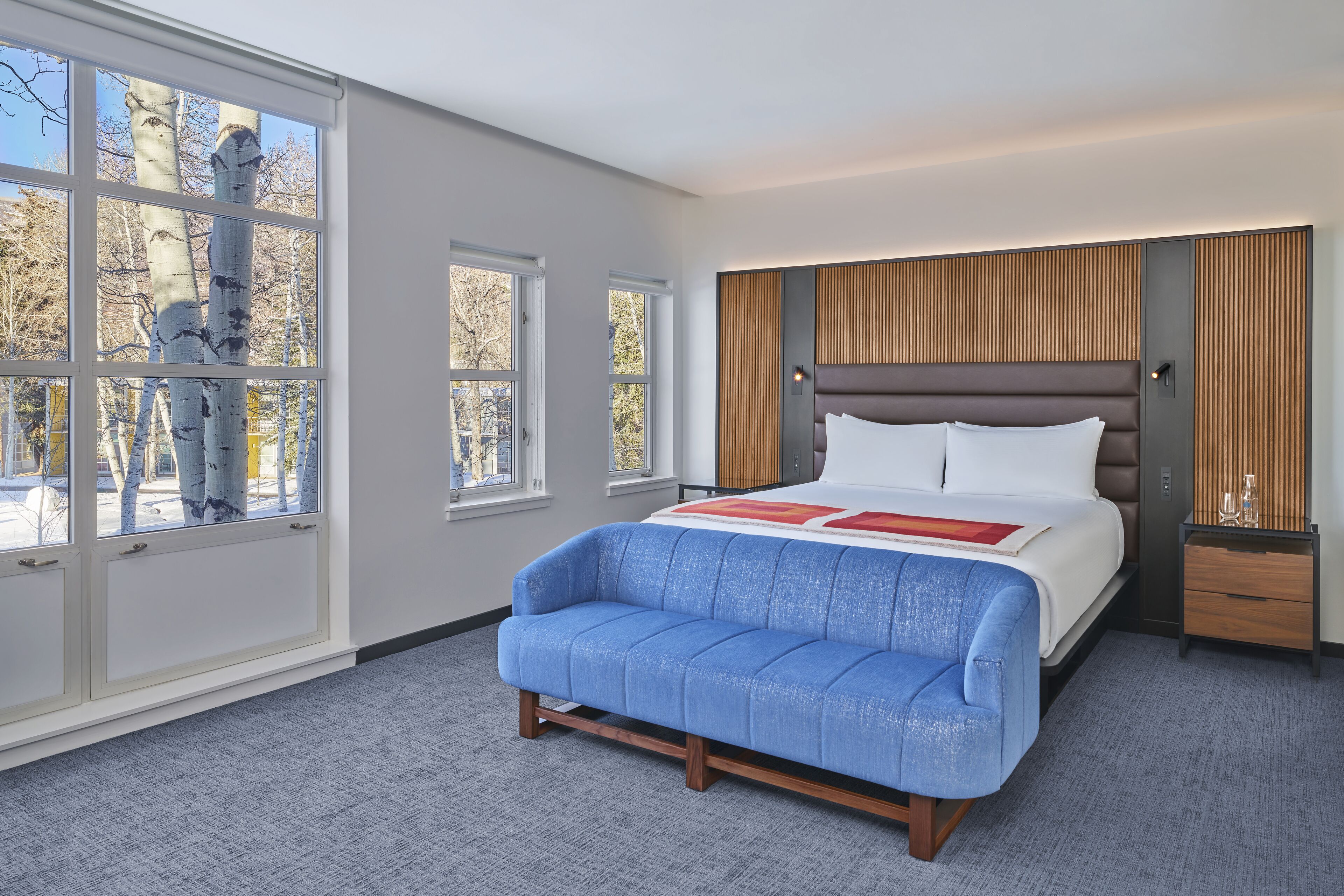Un dormitorio moderno con una cama grande, un sofá azul y vistas a árboles a través de amplios ventanales.