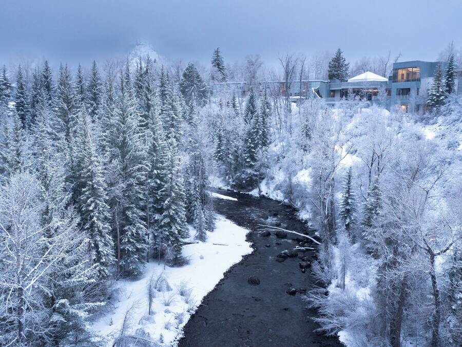 Un bosque cubierto de nieve con un río, y un edificio moderno con varios balcones al fondo.