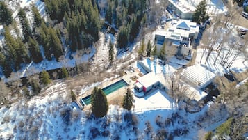 An aerial view of a modern house with a swimming pool, surrounded by snow-covered landscape and dense forest.