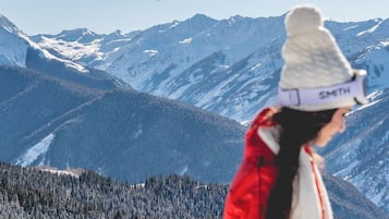 A person in a red and white jacket with a name tag, standing in a mountainous area with snow-covered peaks and a forested valley.