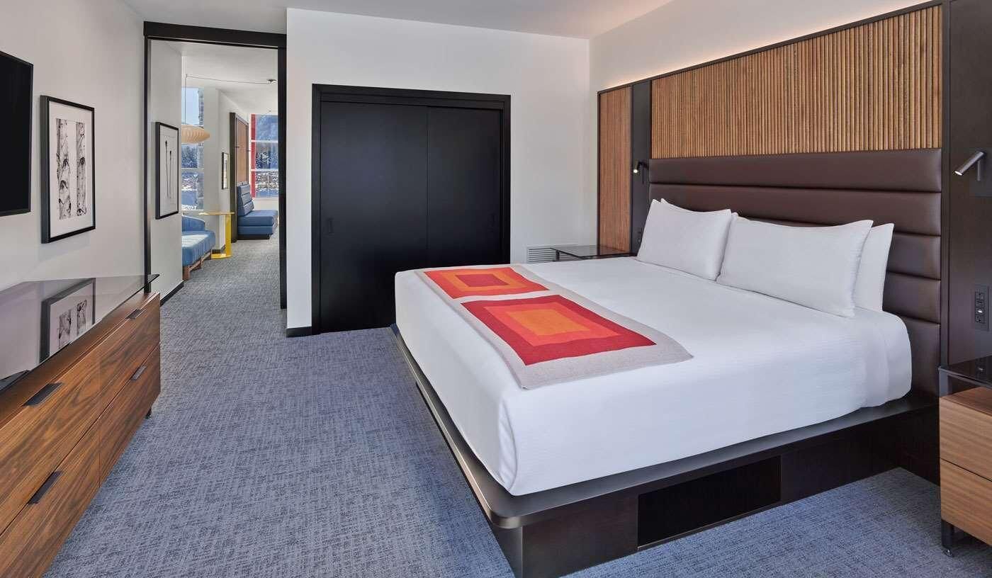 A modern hotel room with a large bed, a wooden headboard, a desk, and a view of the corridor.