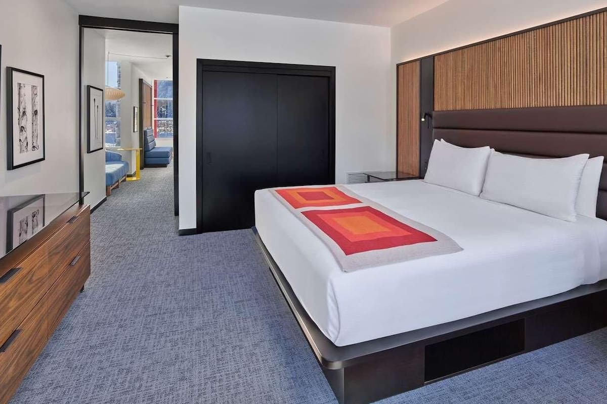 A modern hotel room with a large bed, a wooden headboard, a desk, and a view of the corridor.