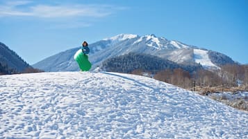 A snowy landscape with a person in a green jacket and helmet, a clear blue sky, and snow-covered mountains in the background.