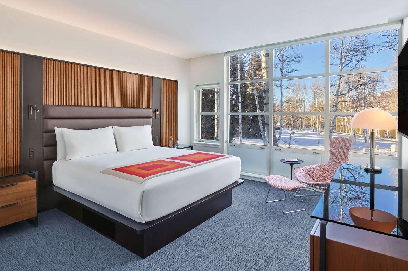 A modern hotel room with a large bed, a desk, a chair, and a view of trees through large windows.