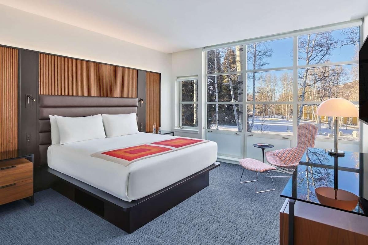 A modern hotel room with a large bed, a desk, a chair, and a view of trees through large windows.