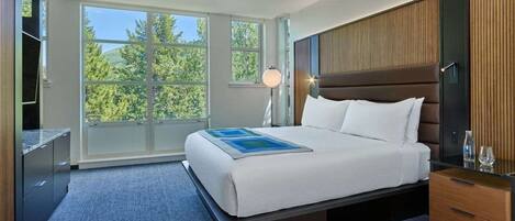 Een moderne hotelkamer met een groot bed, een nachtkastje, een bureau en uitzicht op bomen door grote ramen.