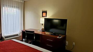 A hotel room with a bed, a desk with a television, a chair, a lamp, and a window with curtains.