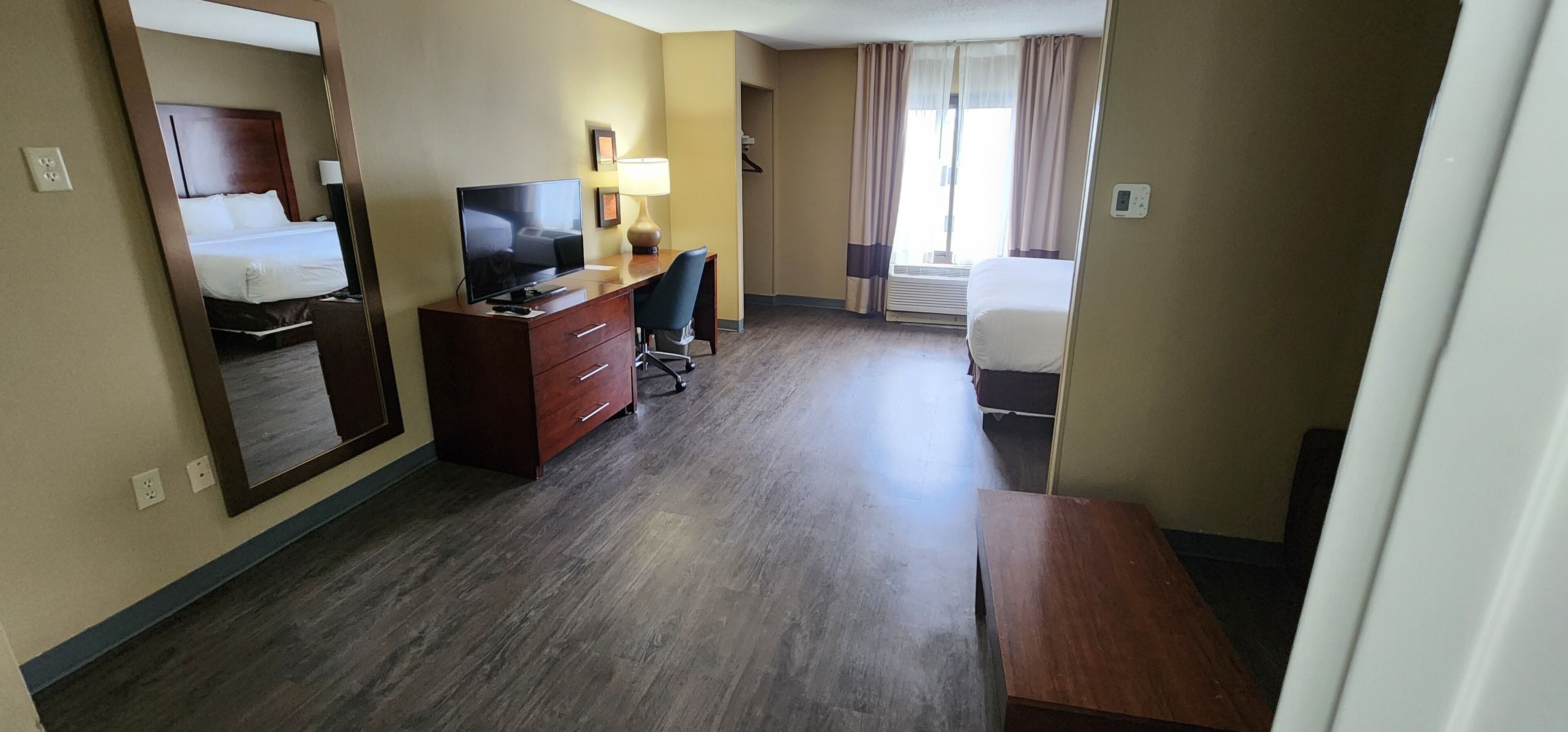 A hotel room with a bed, a desk with a TV, a chair, a lamp, and a window with curtains.