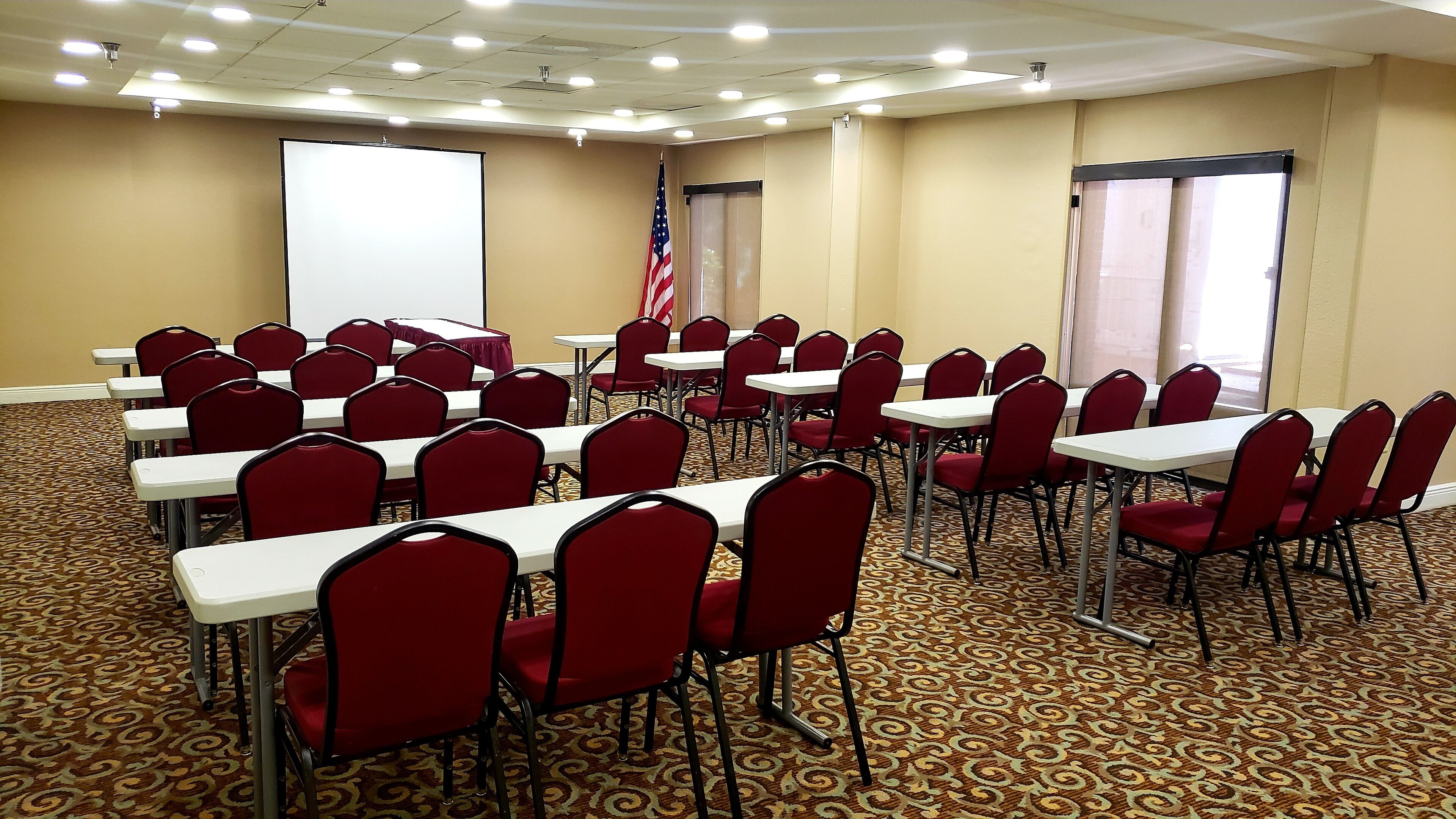 A conference room with rows of tables and chairs, a projector screen, an American flag, and a patterned carpet.