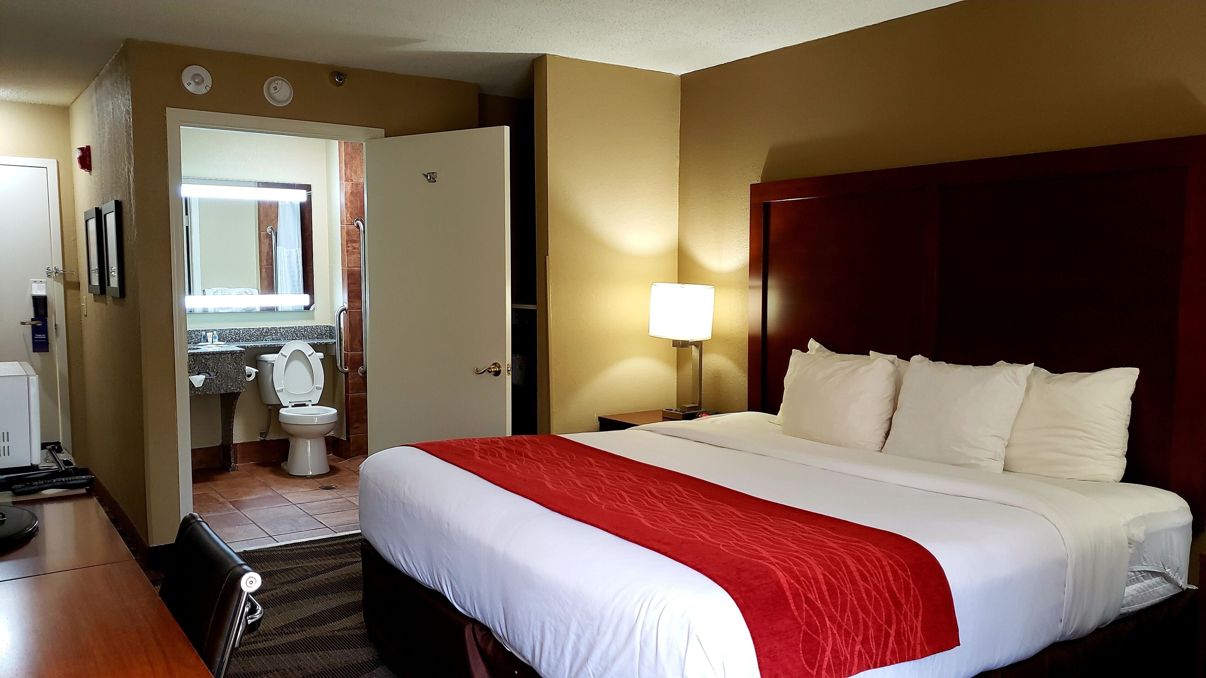A hotel room with a bed, a desk, a chair, a lamp, and a bathroom with a toilet and sink.