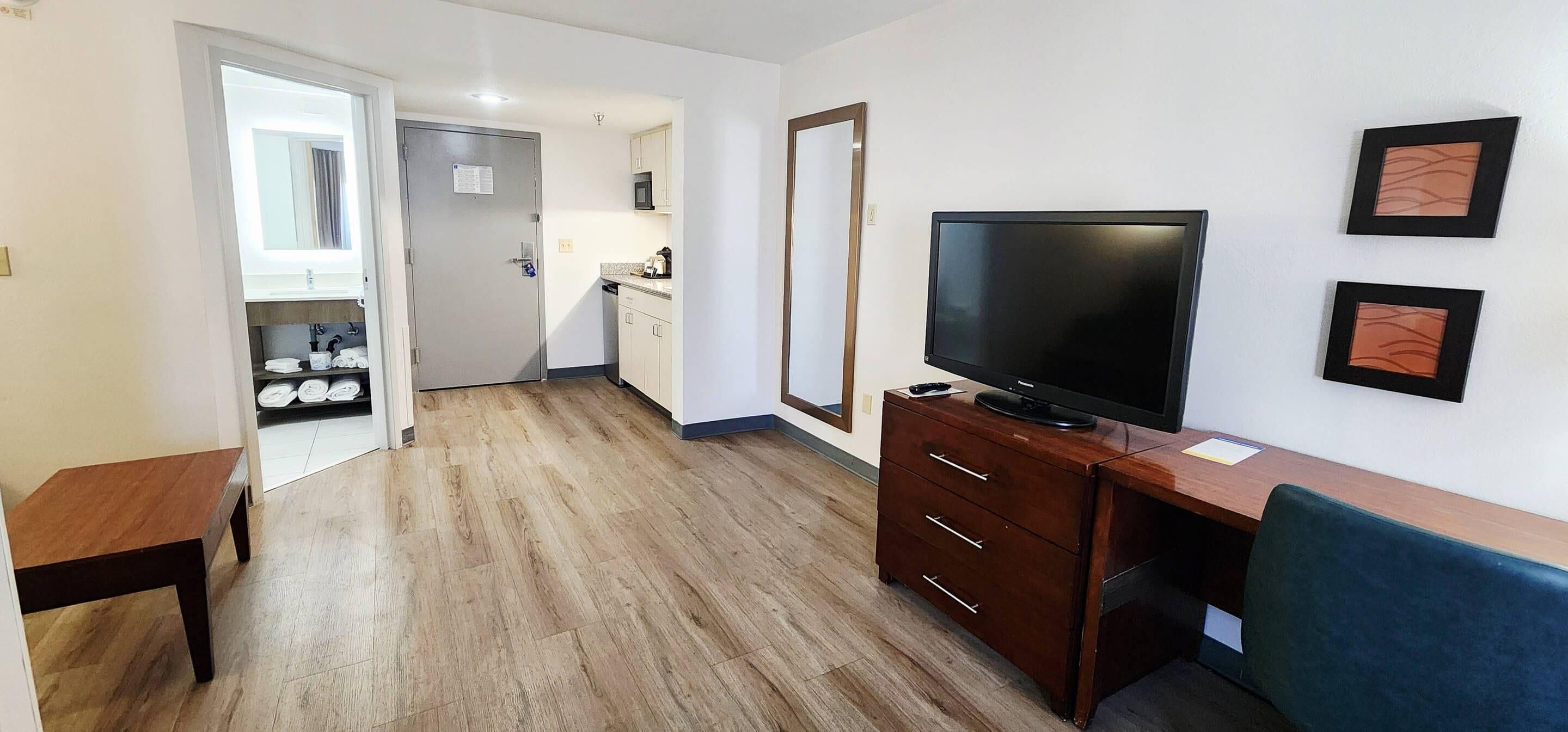 Une pièce avec un plancher en bois, un téléviseur posé sur un meuble en bois, un bureau avec une chaise, un miroir et, au fond, une petite cuisine.