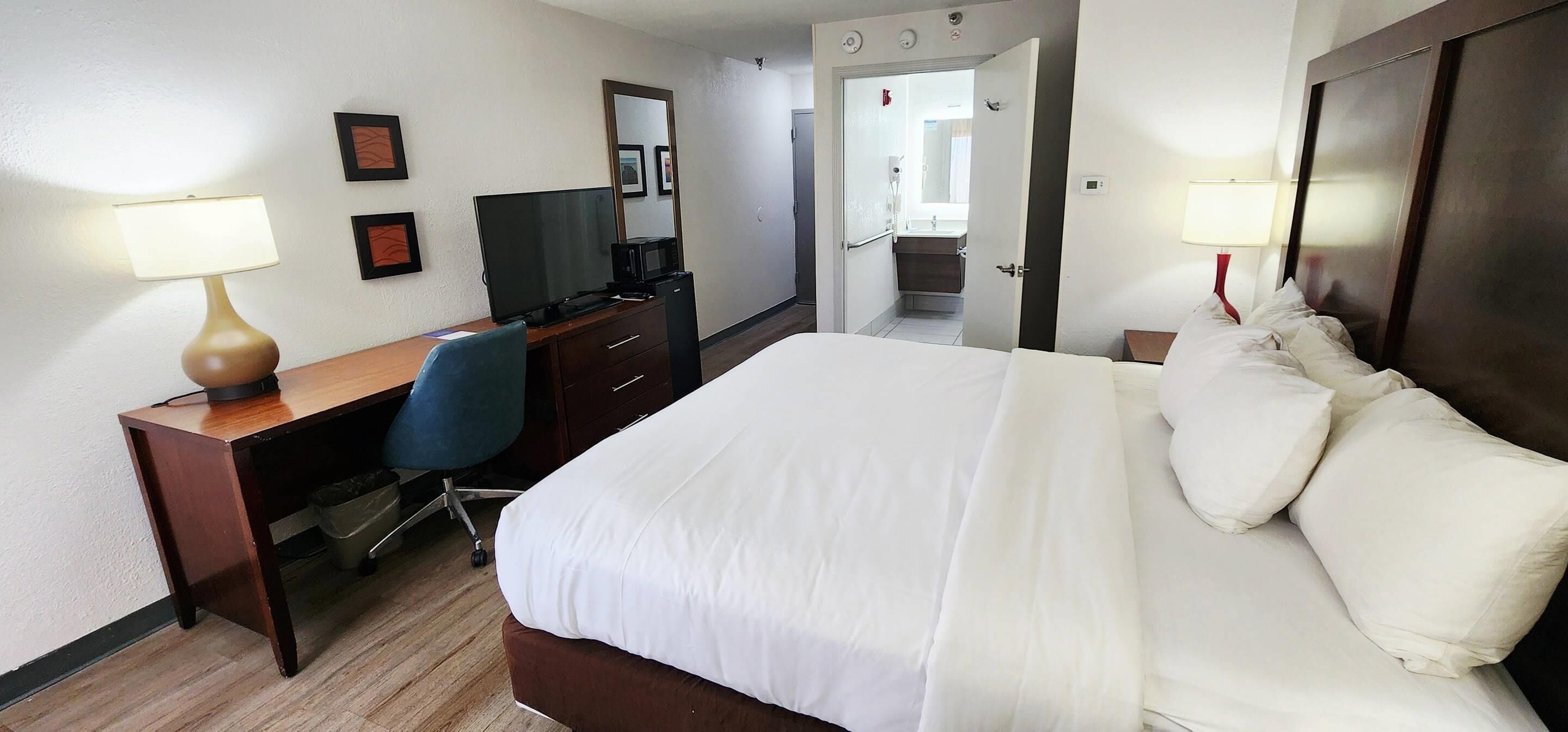 Habitación de hotel con cama, escritorio, silla, televisor y un baño visible a través de una puerta abierta.