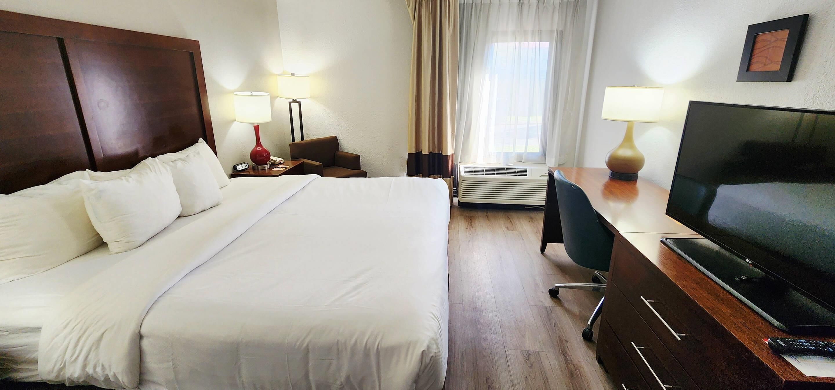 A hotel room with a large bed, a desk with a TV, a chair, an air conditioning unit, and a window with curtains.