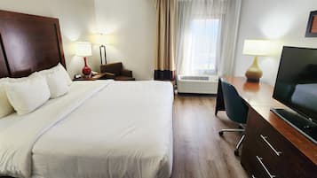 A hotel room with a large bed, a desk with a TV, a chair, an air conditioning unit, and a window with curtains.