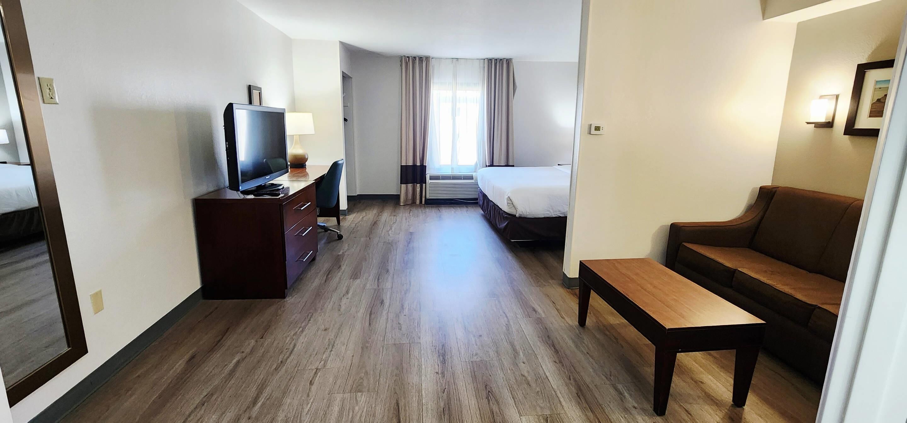 Een hotelkamer met een houten vloer, een televisie op een bureau, een bank, een bed en een raam met gordijnen.