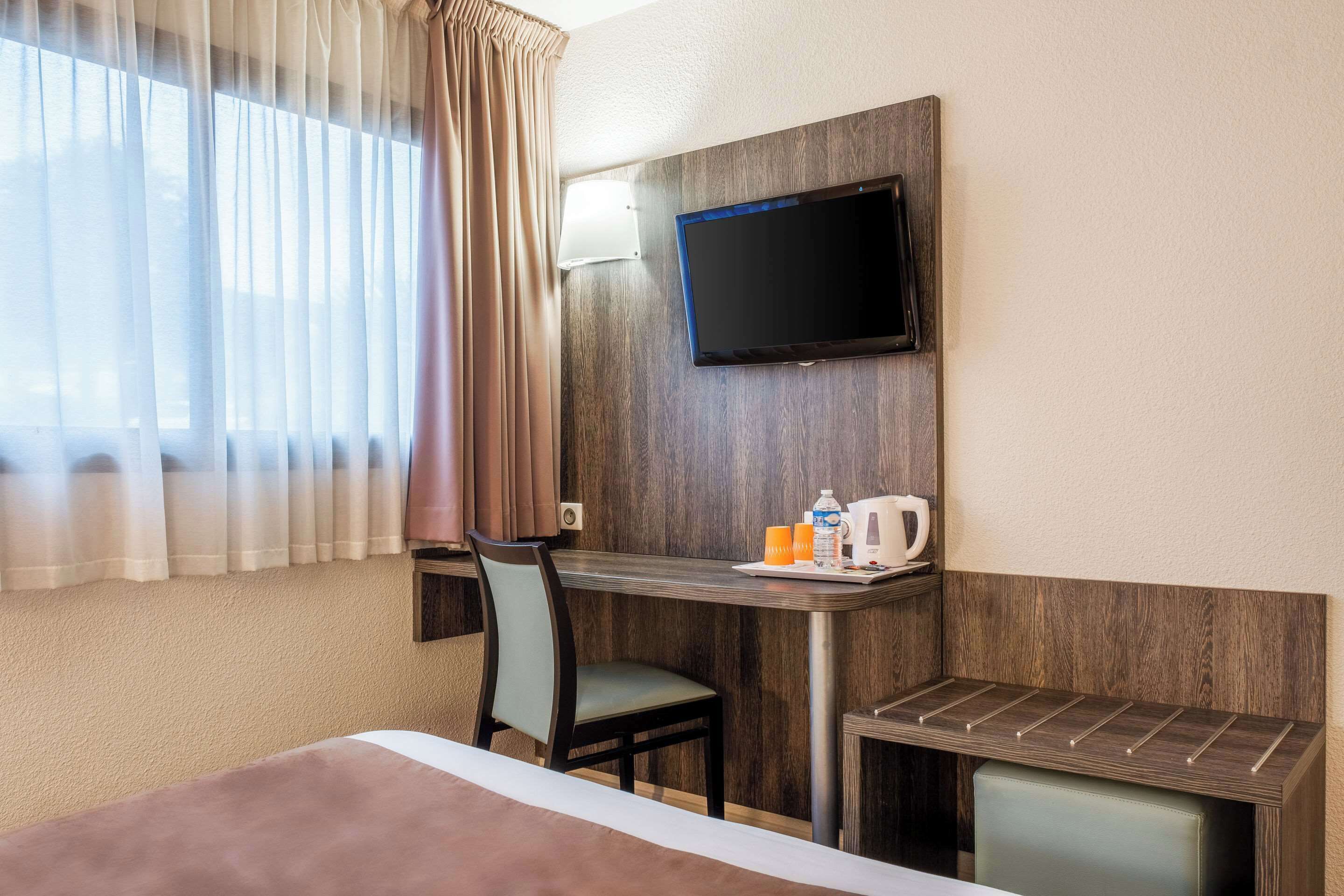 A hotel room with a bed, a desk with a chair, a television mounted on the wall, and a window with curtains.