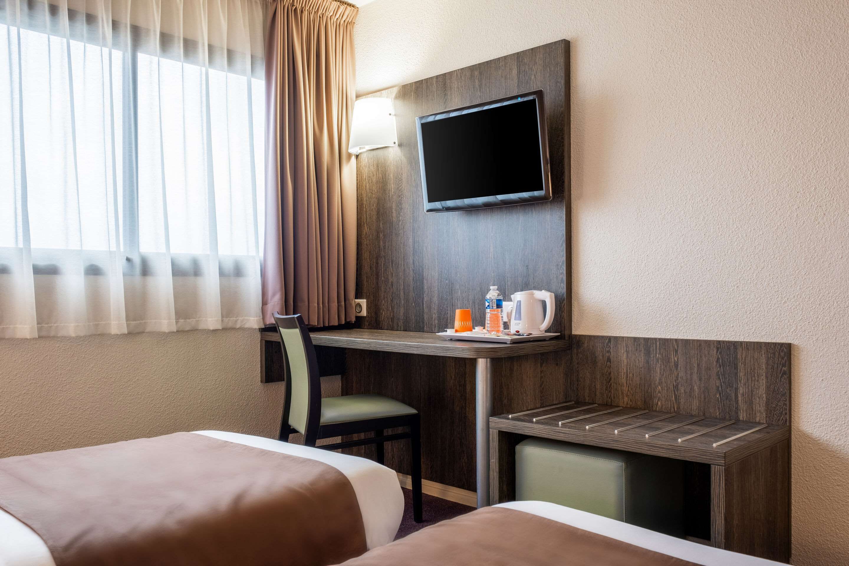 A hotel room with two beds, a desk with a flat-screen TV, a chair, and a small table with a kettle and cups.