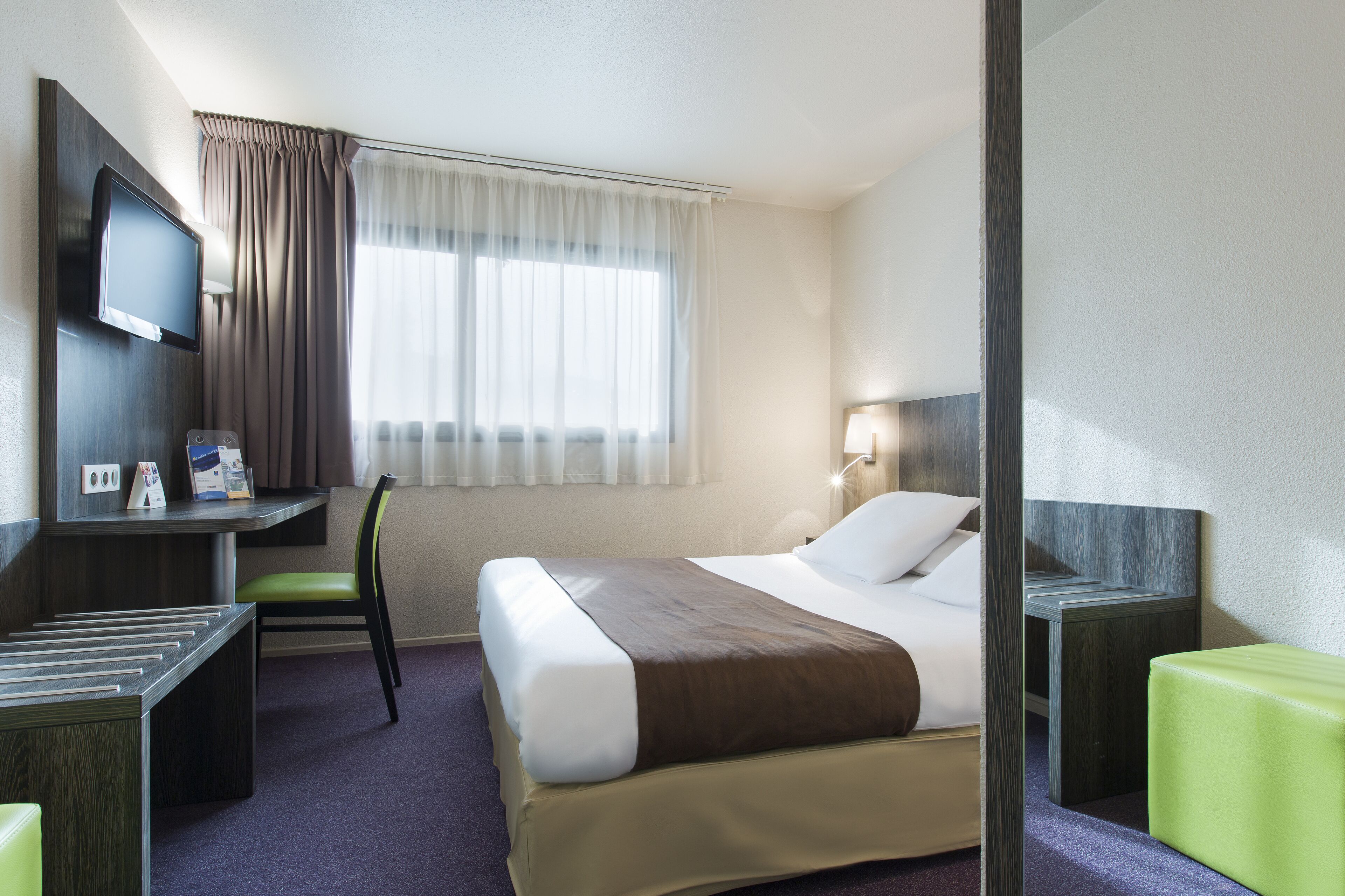Een hotelkamer met een bed, een bureau met stoel, een televisie en een raam met gordijnen.