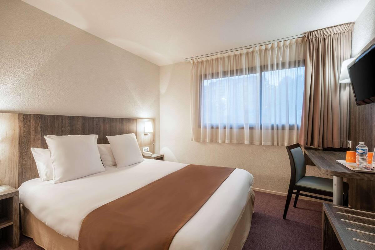 Ein Hotelzimmer mit einem großen Bett, einem Schreibtisch mit Stuhl, einem Fernseher und einem Fenster mit Vorhängen.