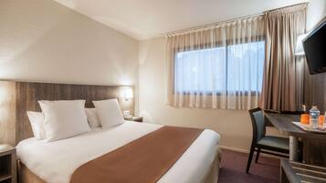 Ein Hotelzimmer mit einem großen Bett, einem Schreibtisch mit Stuhl, einem Fernseher und einem Fenster mit Vorhängen.