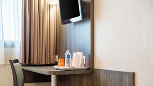 Une chambre d’hôtel avec un bureau en bois, une chaise, un téléviseur à écran plat fixé au mur, une bouteille d’eau, une machine à café et un verre posé sur le bureau.