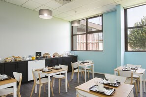 Un espace repas moderne avec des tables blanches et en bois, des meubles noirs et de grandes fenêtres qui laissent entrer la lumière naturelle.