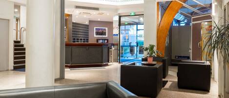 A modern hotel lobby with a reception desk, seating areas, and a view of the street.
