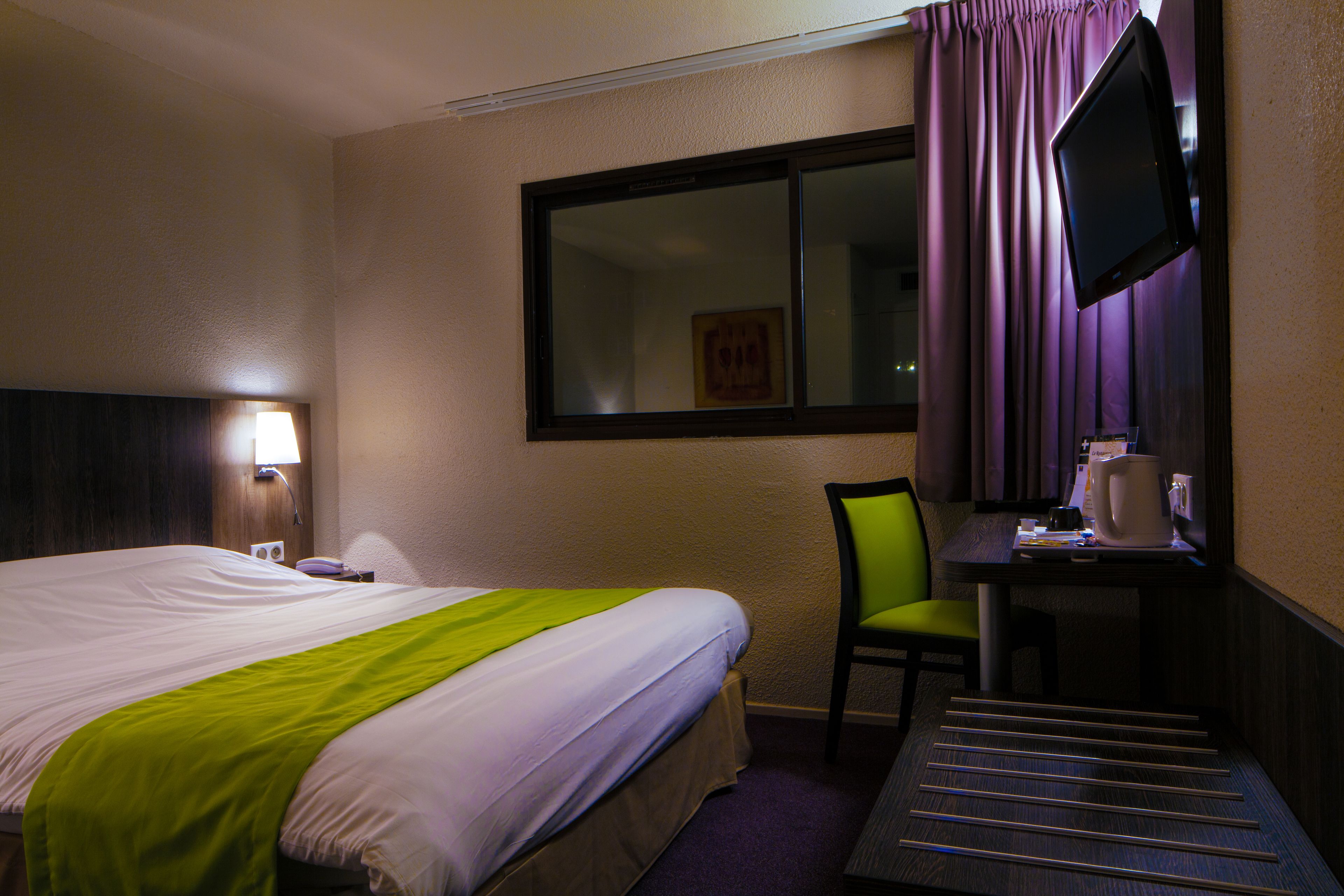 Habitación de hotel con cama, escritorio, silla, televisor y ventana con cortinas.