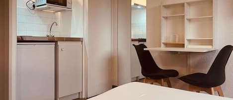 A compact kitchen with a microwave, a sink, and a small refrigerator. There is a dining area with a white table and two black chairs.
