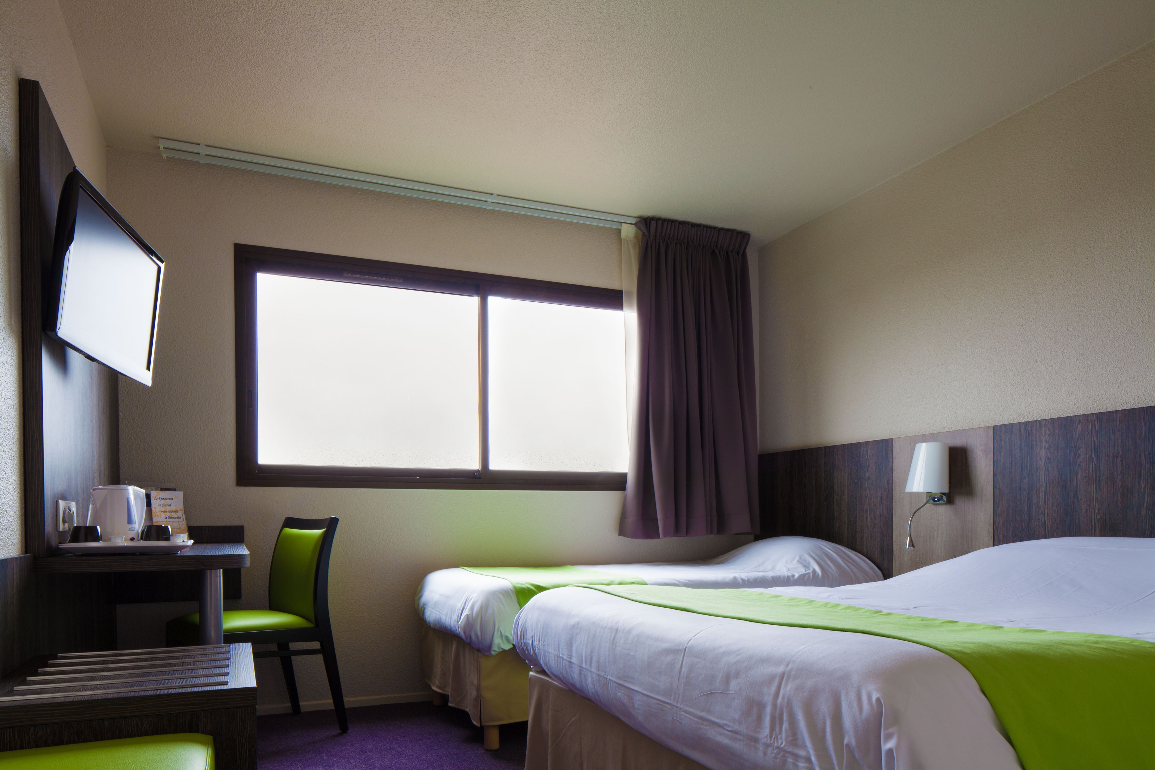 Ein Hotelzimmer mit zwei Betten, einem Schreibtisch mit Stuhl, einem Fernseher und einem Fenster mit Vorhängen.