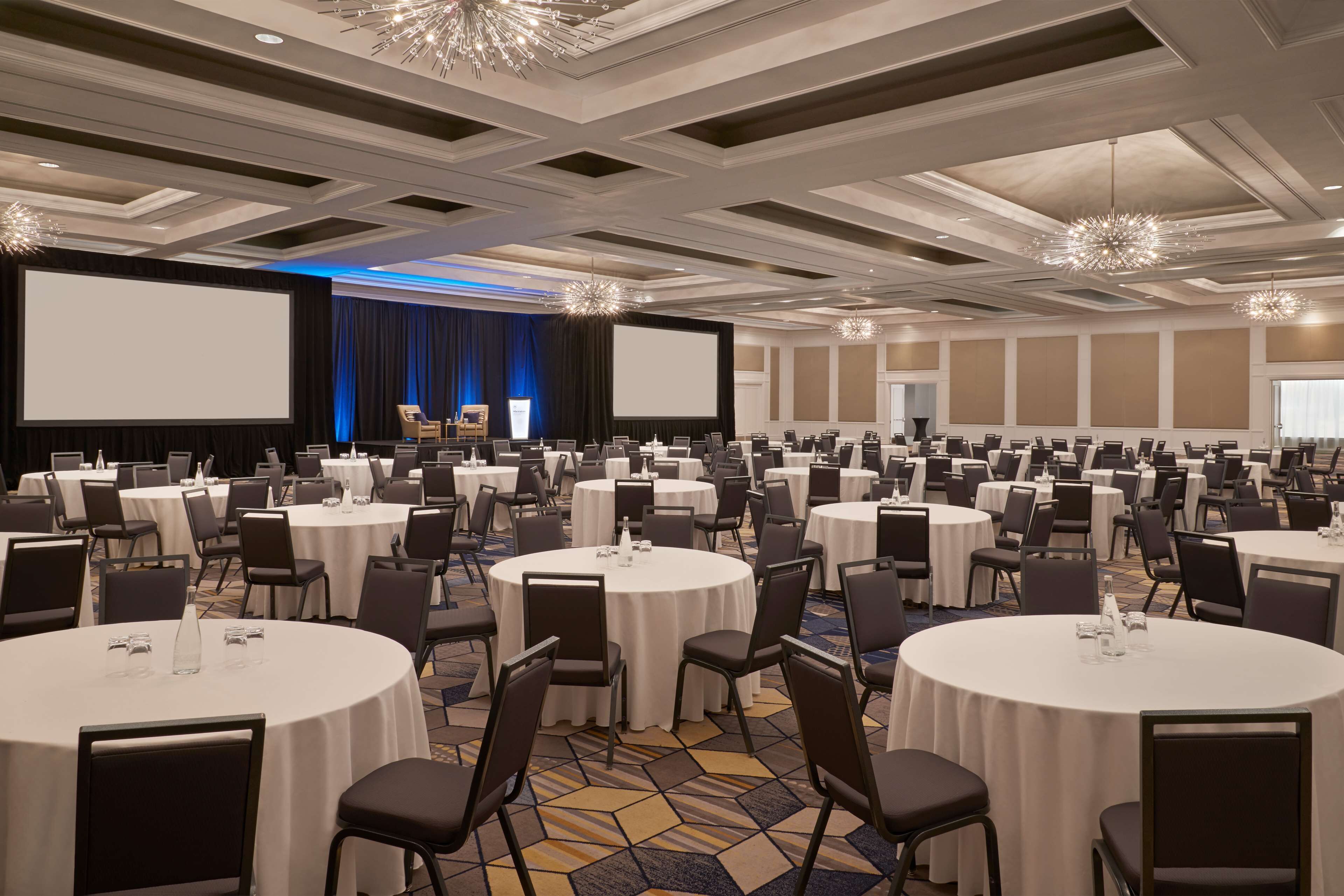 A large conference room with multiple round tables, a stage with a blue curtain, and a large screen.