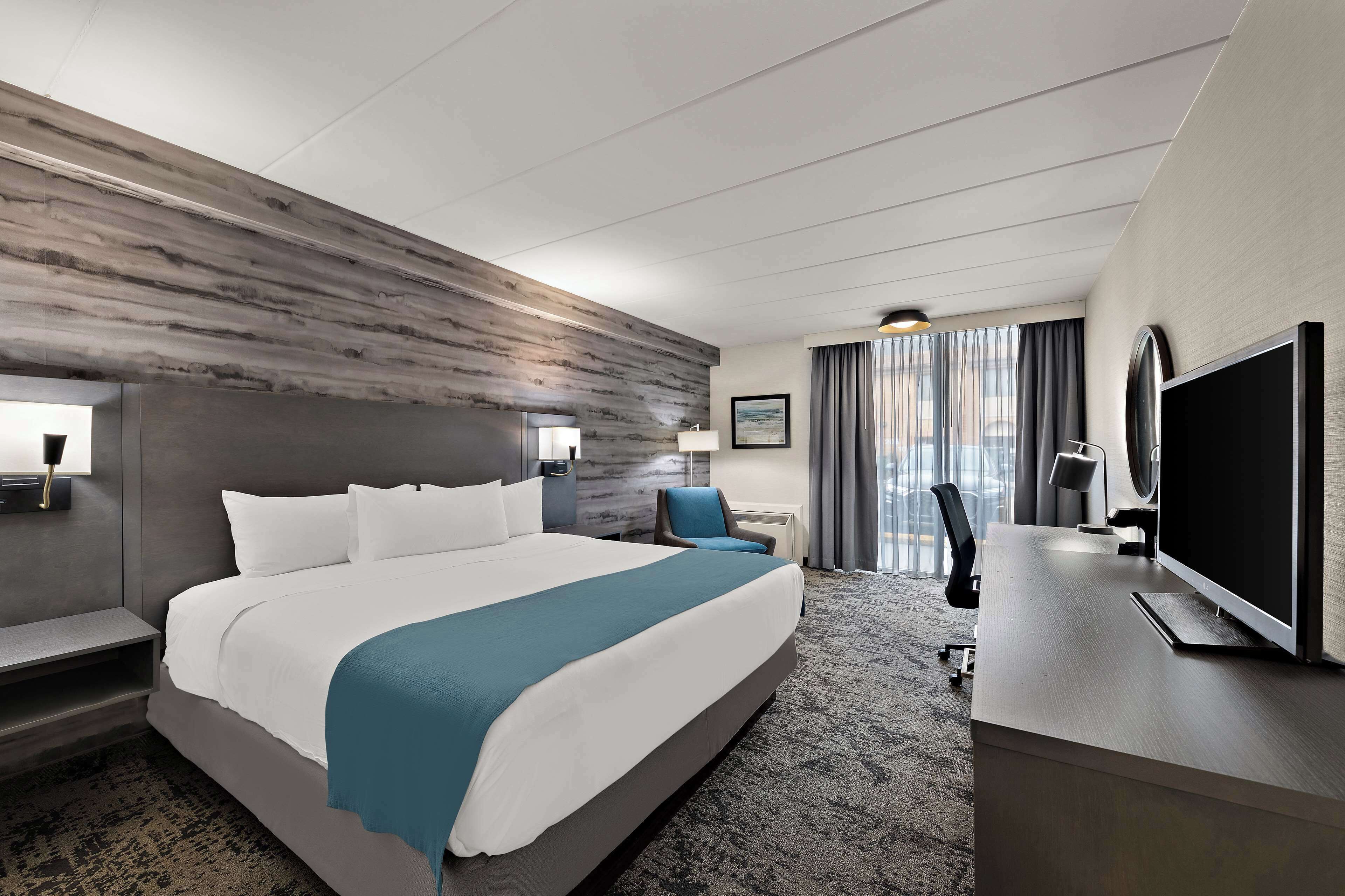 Una habitación de hotel moderna con una cama grande, un escritorio, un televisor de pantalla plana, una silla y un ventanal con cortinas.
