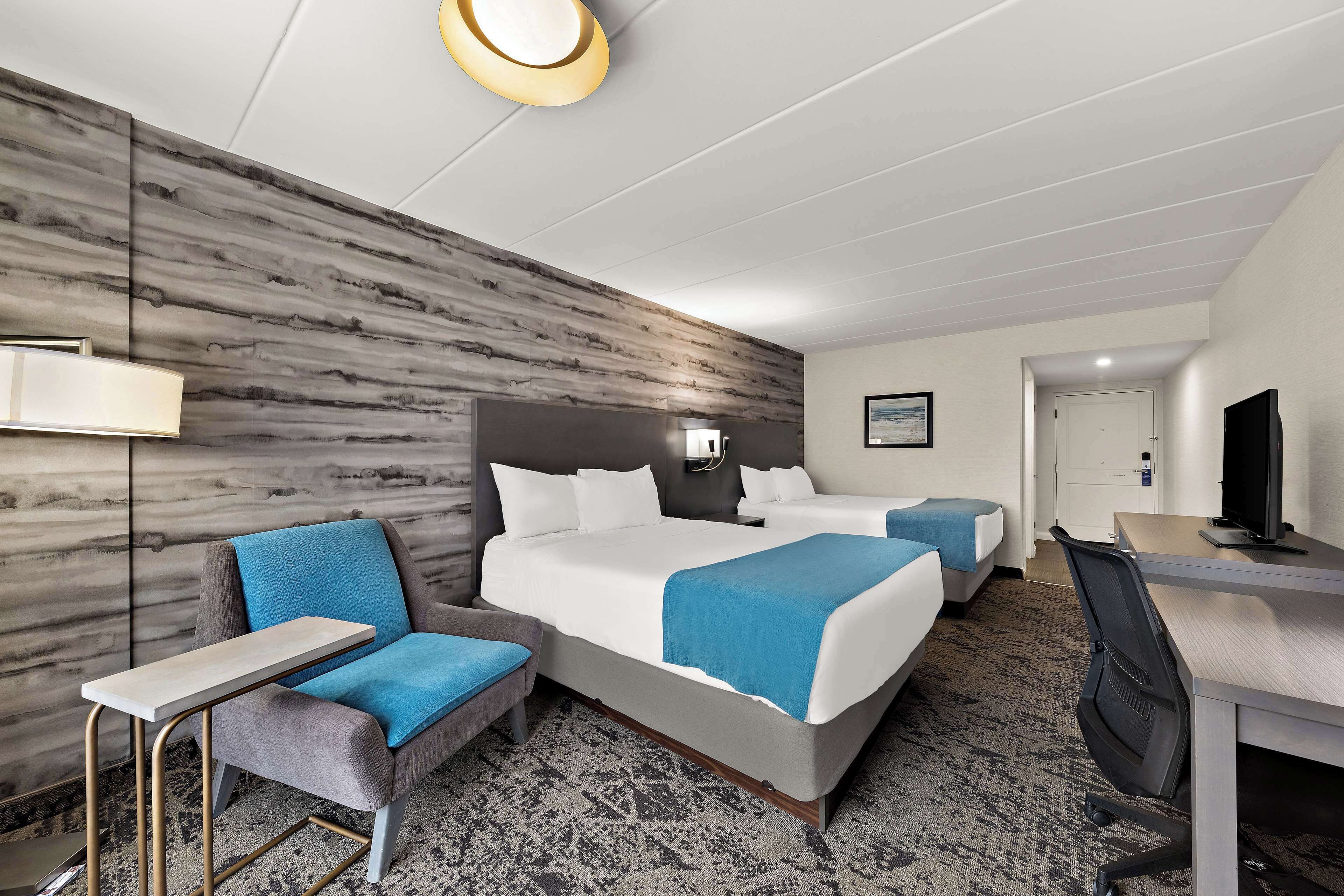 A hotel room with two beds, a desk, a chair, a lamp, and a television.