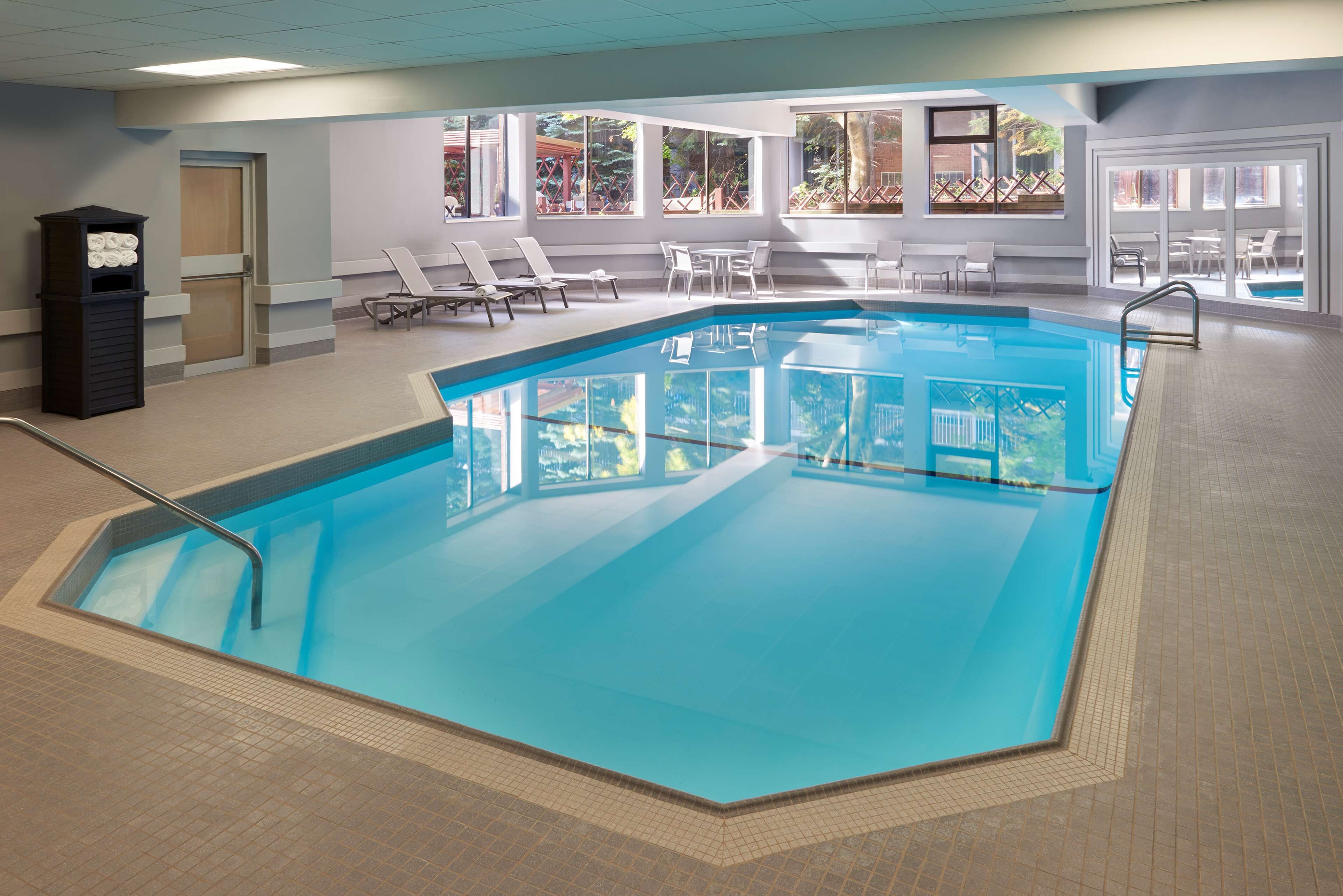 A large indoor swimming pool with clear blue water, surrounded by lounge chairs and tables, and large windows offering views of the outdoors.