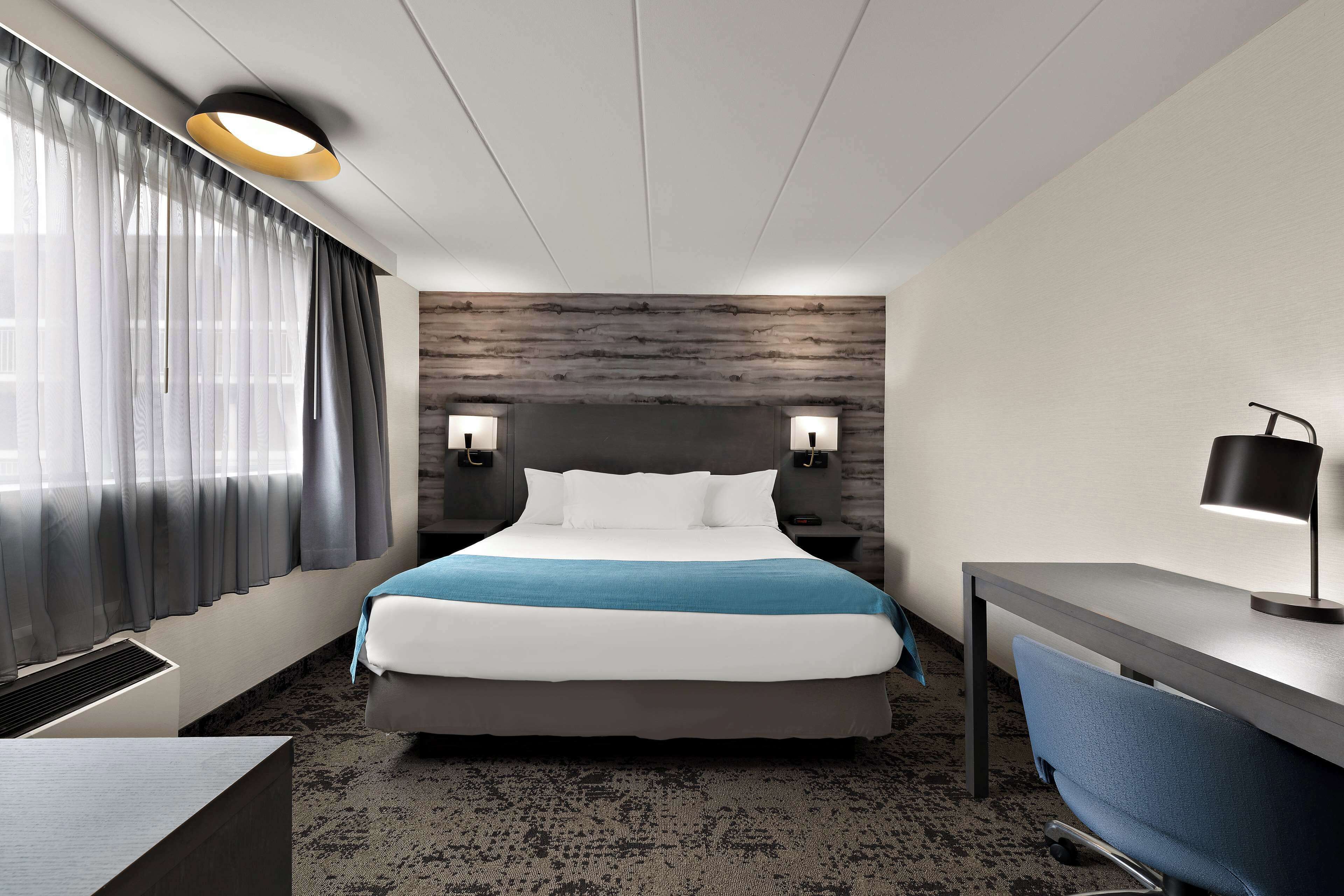 Habitación de hotel moderna con una cama grande, un escritorio y una silla. Hay dos lámparas de pared y una luz de techo.