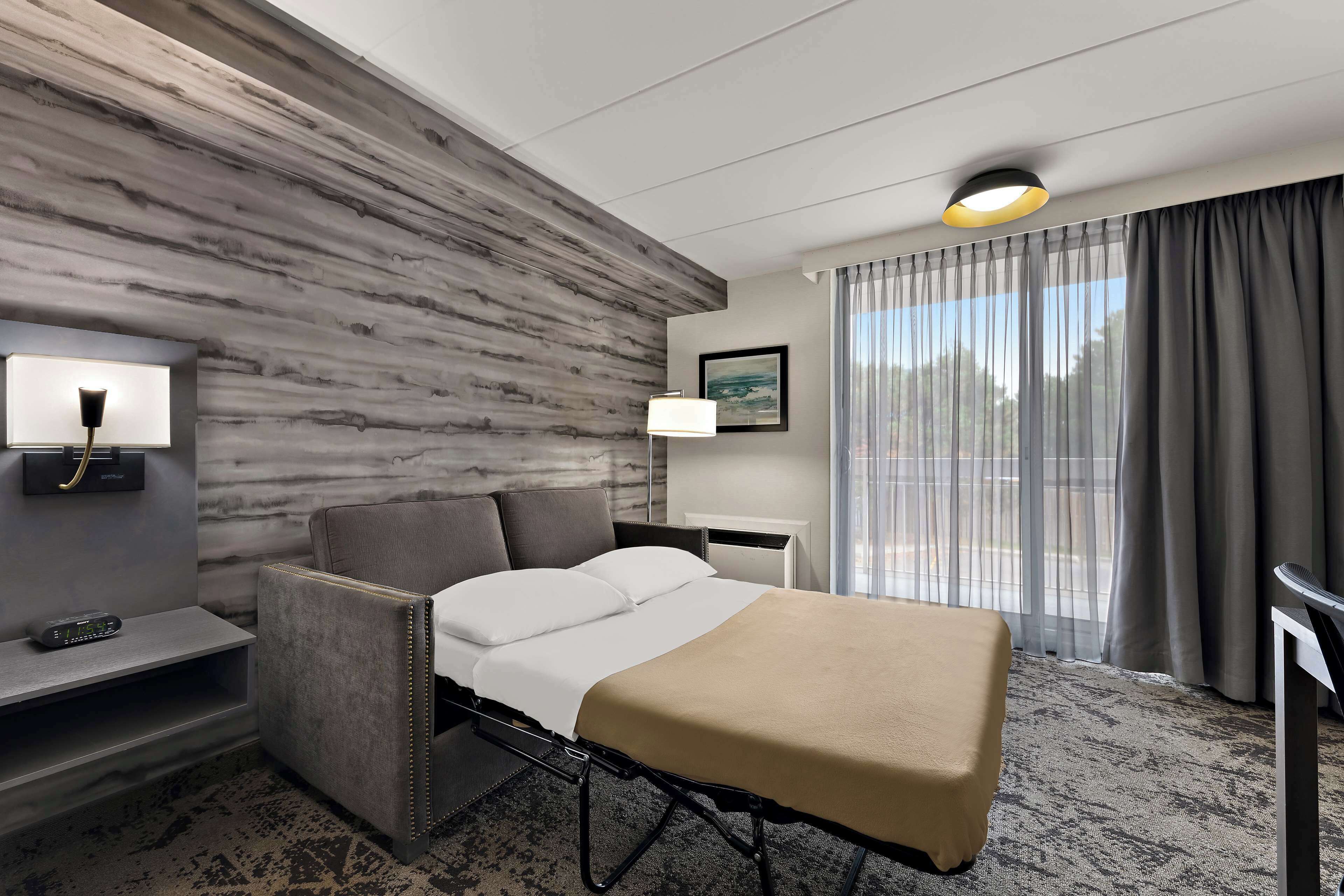 A hotel room with a bed, a desk, a chair, a lamp, and a window with curtains.