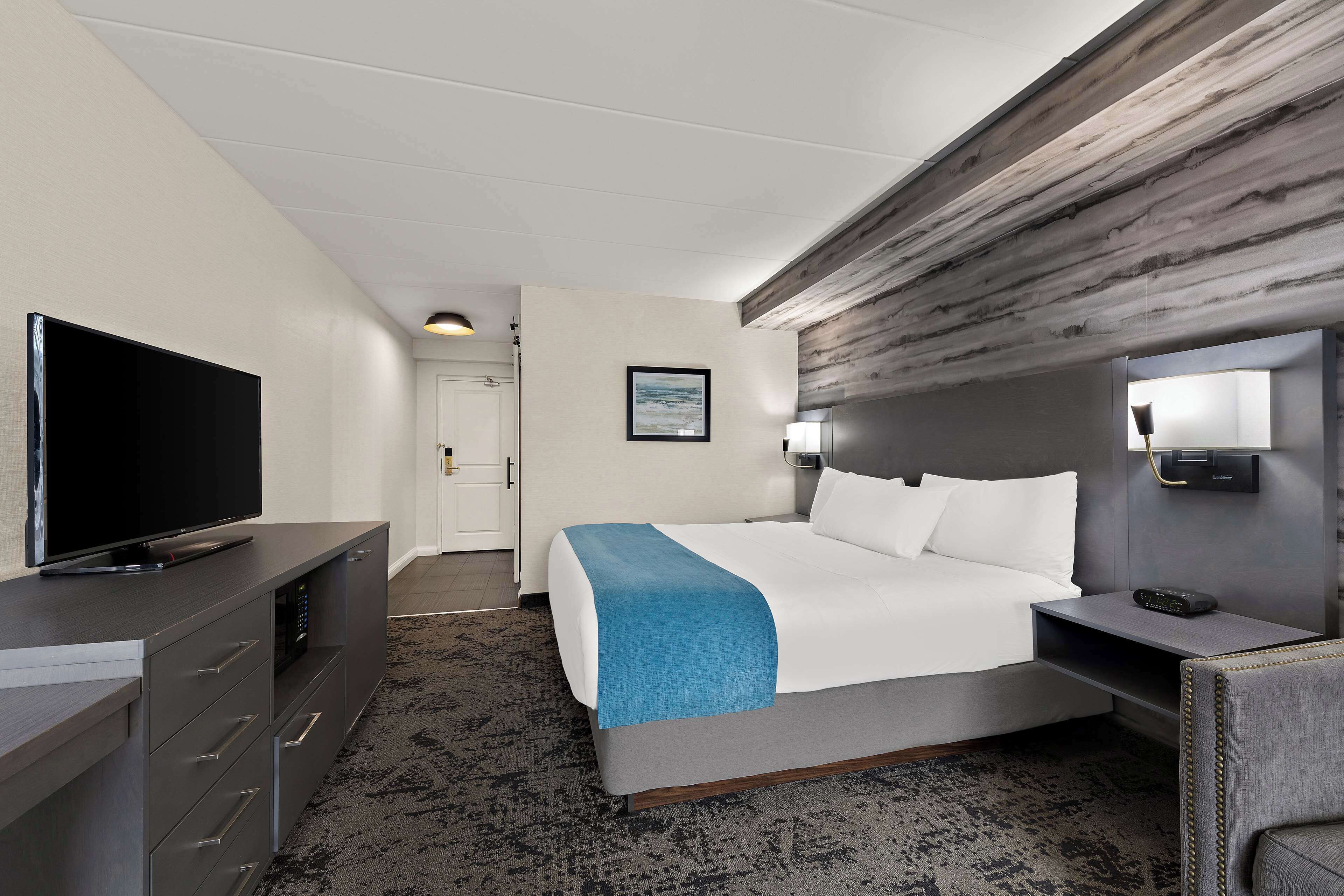 Habitación de hotel con cama, televisor en una cómoda, escritorio con lámpara y un cuadro en la pared.