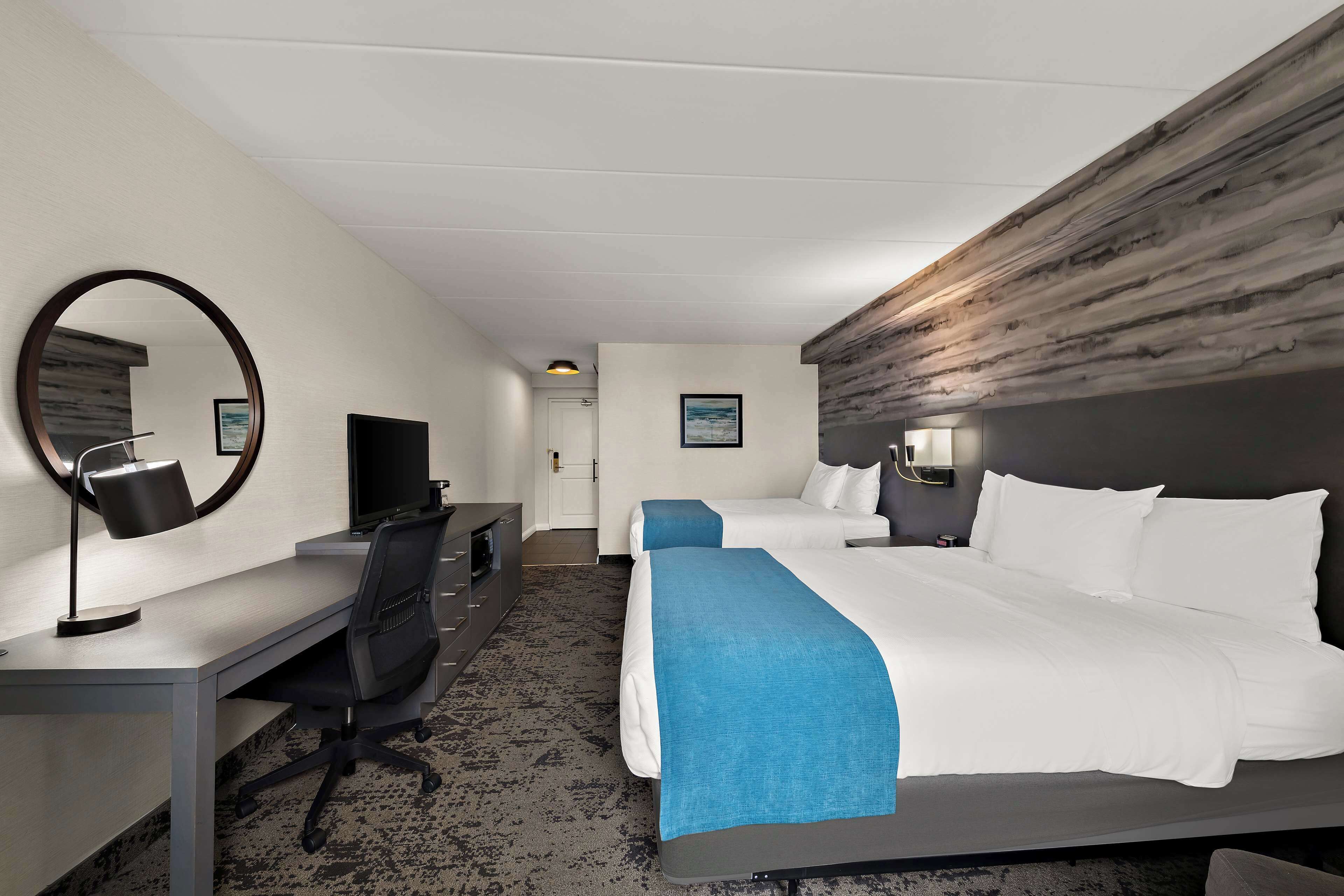 Una habitación de hotel con escritorio, dos camas, televisor y un espejo grande.