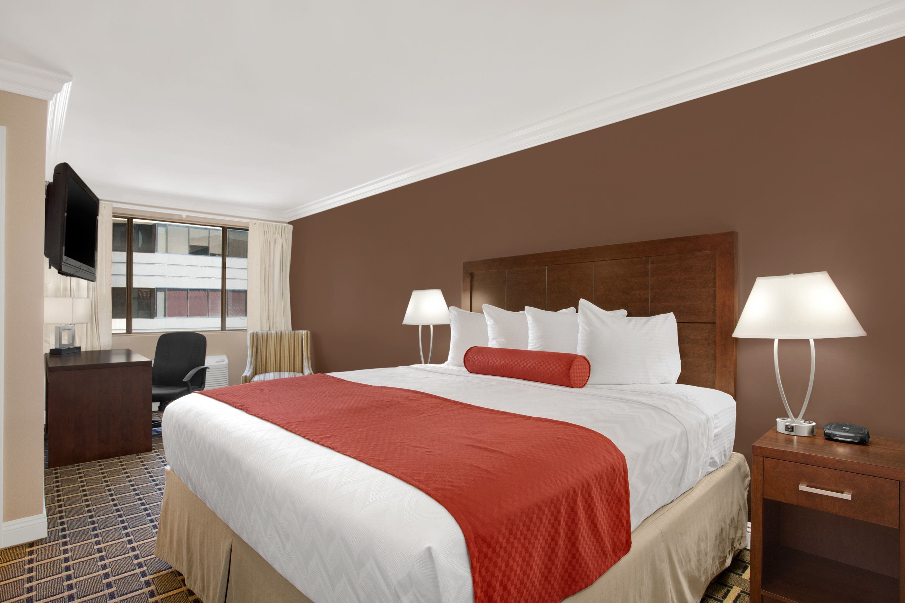 A hotel room with a large bed, two bedside lamps, a wooden headboard, a desk with a chair, and a TV mounted on the wall.