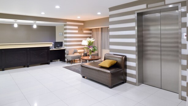 A modern hotel lobby with a reception desk, a sofa with a yellow pillow, a small table with a vase of flowers, and a large elevator. - Best Western Plus LA Mid Town Hotel (Los Angeles)