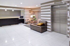 A modern hotel lobby with a reception desk, a sofa with a yellow pillow, a small table with a vase of flowers, and a large elevator. - Best Western Plus LA Mid Town Hotel (Los Angeles)