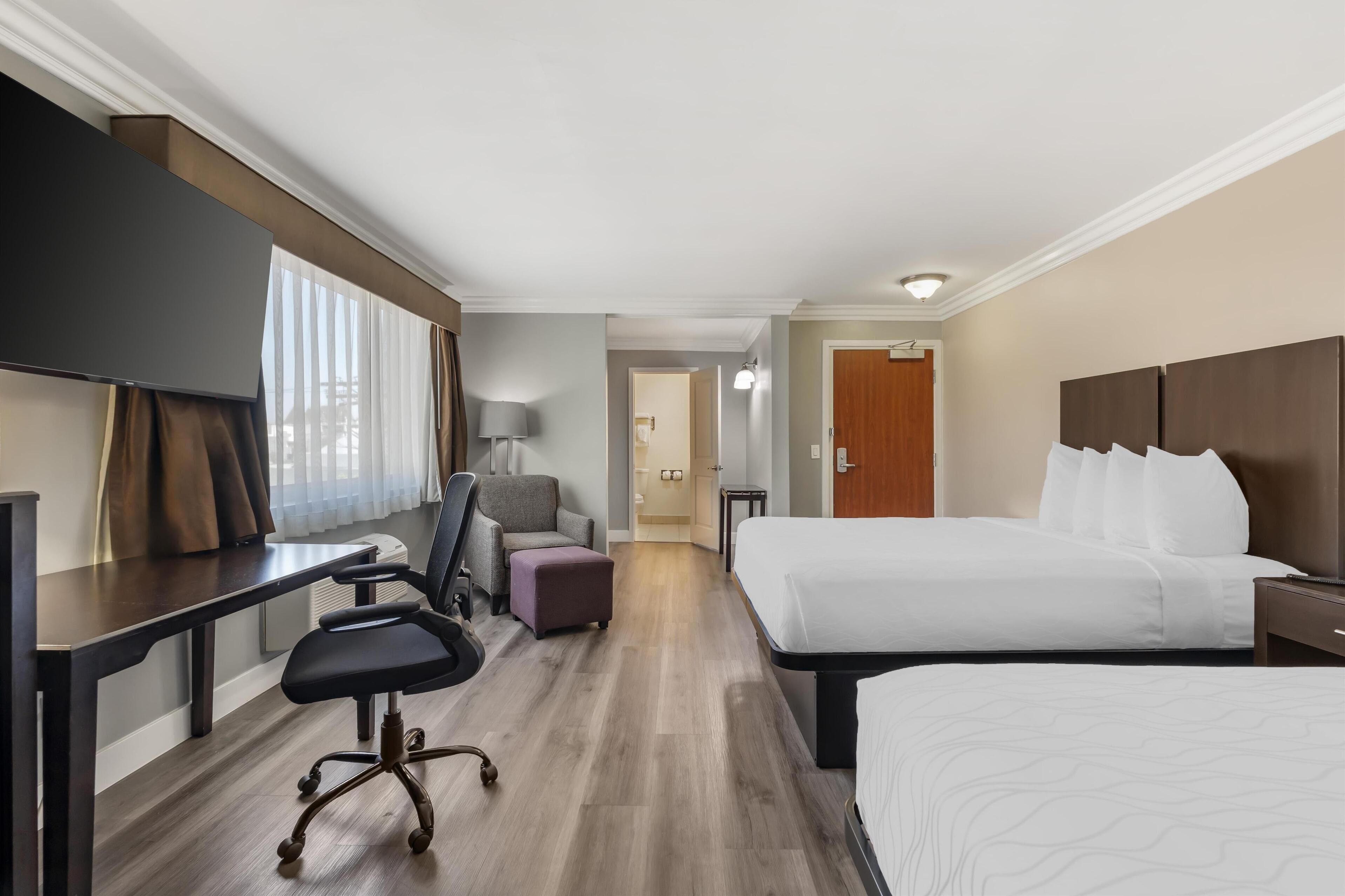 Quarto de hotel com cama, escrivaninha, cadeira, abajur e televisão.