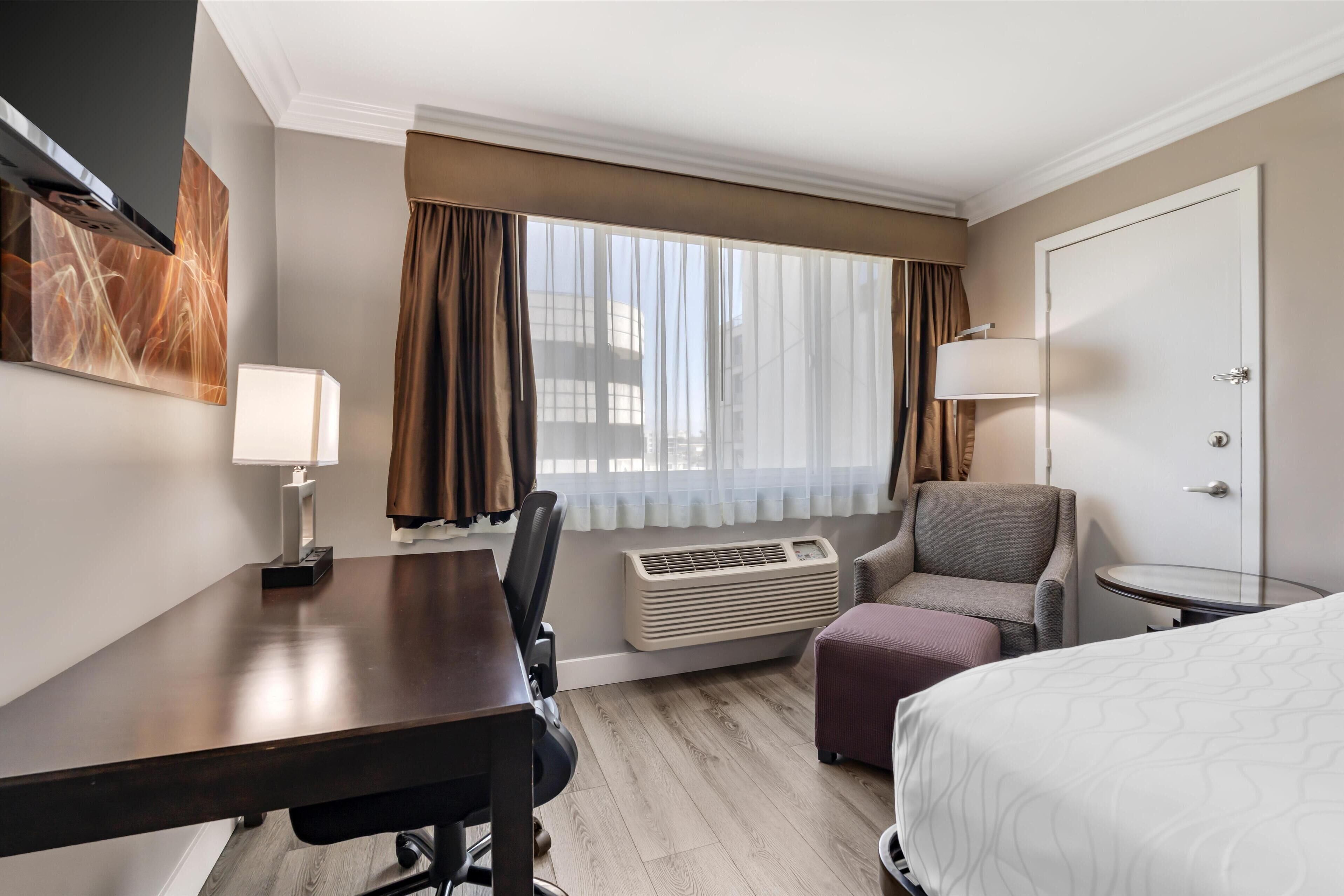 Habitación de hotel con cama, escritorio con lámpara, una silla, una mesita y aire acondicionado.